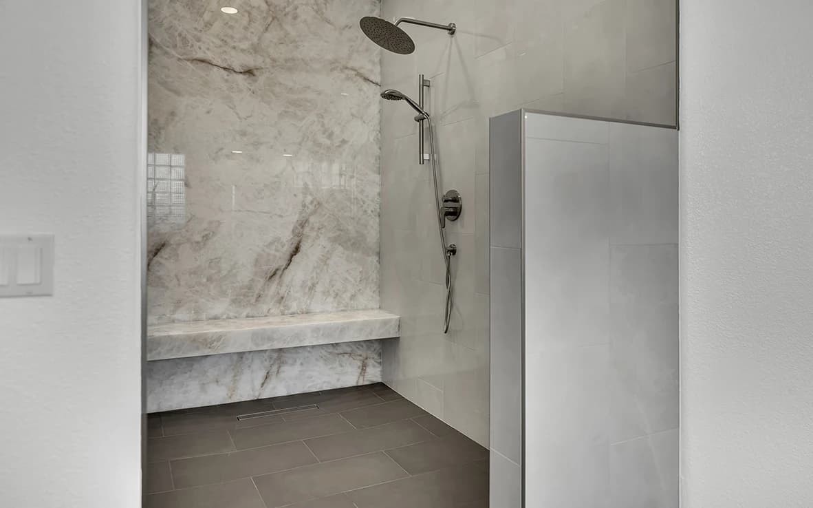 Curbless primary bathroom remodel with Honey Amber Onyx slab shower wall, bench, linear drain, rain head, handheld wand, and charcoal gray tile floor